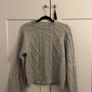 Grå stickad tröja med flätmönster - Mysig grå stickad tröja med klassiskt flätmönster och rund hals. Tröjan har långa ärmar och ribbade muddar vid ärmslut och nederkant. Enkel att matcha med olika outfits.