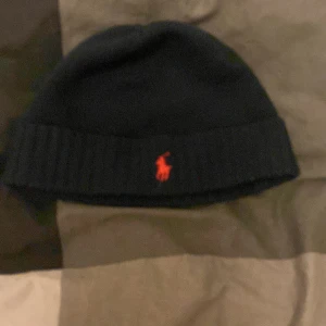 Svart stickad mössa från Polo Ralph Lauren - Svart stickad mössa från Polo Ralph Lauren med röd broderad logga framtill. Klassisk ribbad kant och mjukt material som håller värmen. Perfekt accessoar för kyliga dagar.