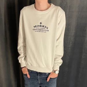 Vit sweatshirt från Morris - Säljer en stilren vit sweatshirt från Morris med tryckt logga och text på bröstet. Tröjan har rund halsringning och ribbade muddar vid ärmslut och nederkant. Perfekt för en avslappnad och snygg look. Tröjan är liten i storleken så passar mer en M. Skick 8/10✅ Pris kan diskuteras vid snabb affär💸