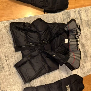 Svart dunjacka med huva från Burberry - Säljer en svart dunjacka från Burberry med klassiskt rutigt foder i huvan. Jackan har dragkedja, knappar och två fickor framtill. Perfekt för kyliga dagar och har en snygg, tidlös design.