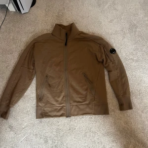 Beige jacka från C.P. Company - Säljer en beige tröja från C.P. Company med hög krage och dragkedja framtill. Tröjan har två snedställda fickor med dragkedja och en ikonisk linsdetalj på ärmen. Perfekt för dig som gillar stilrena och funktionella plagg. Vid mer funderingar är det bara att höra av sig
