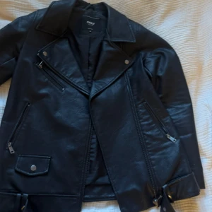 Svart skinnjacka från Only - Svart skinnjacka från Only med klassisk bikerstil. Jackan har sned dragkedja, stora slag, fickor med dragkedja och bälte i midjan.