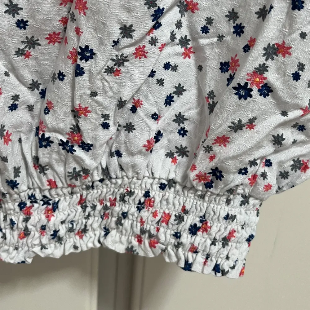Söt vit blus med små blommor i rosa, blått och grått. Blusen har korta puffärmar med resår och resår i midjan för en feminin siluett. Perfekt för vår och sommar, lätt och luftig i bomull.. Yläosat.