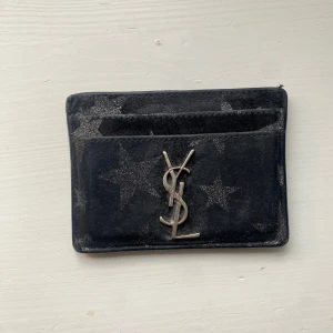 Svart korthållare från Saint Laurent med stjärnor - Säljer en svart korthållare från Saint Laurent med glittrigt stjärnmönster och ikoniska YSL-loggan i silver framtill. Perfekt för kort och småsaker, smidig att ha i fickan eller väskan. Sliten men inte trasig!