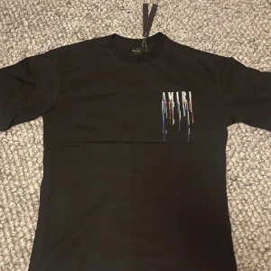 Svart t-shirt från Amiri med färgstänk - Säljer en svart t-shirt från Amiri med tryckt logga och färgstänk i blått, vitt och orange på bröstet och ryggen. Klassisk passform och rund halsringning. Perfekt för dig som gillar streetwear och vill sticka ut.