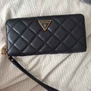 Svart quiltad plånbok från Guess - Snygg svart quiltad plånbok från Guess med guldfärgade detaljer och dragkedja runtom. Plånboken har ett klassiskt rutmönster och en praktisk handledsrem. Perfekt för att hålla ordning på kort och kontanter.