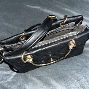 Svart quiltad handväska med gulddetaljer - Snygg svart handväska med quiltat mönster och guldfärgade handtag. Väskan har dragkedja upptill och både axelrem och handtag i metall. Perfekt för dig som vill ha en stilren accessoar med lyxig känsla.