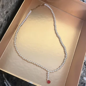 Pärlhalsband med röd detalj - Elegant halsband med vita pärlor och en liten röd pärldetalj längst ner. Kedjan är i guldton och halsbandet har ett klassiskt och stilrent utseende, perfekt för att lyfta vilken outfit som helst.