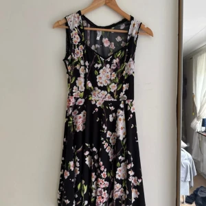 Blommig svart klänning Chiara Forti - Supersöt svart klänning från Chiara Forti med v-ringning och blommigt mönster i vitt, rosa och grönt. Ärmlös modell med markerad midja och lätt utställd kjol. Perfekt för sommarens alla tillfällen!