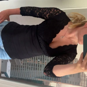 Svart spetsblus med v-ringning - Supersnygg svart blus med djup v-ringning och trekvartsärmar i spets. Toppen har figurnära passform och vackra spetsdetaljer över axlar, rygg och ärmar. Skitsnygg till fest i andra fina saker att gå på. Aldrig använd