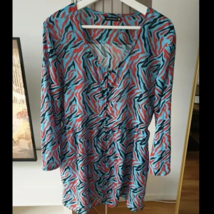 Zebramönstrad kortklänning från Rut&Circle - Färgstark kortklänning från Rut&Circle med blått, rött och svart zebramönster. Klänningen har v-ringning, knappar framtill och långa ärmar. Skön och luftig passform.