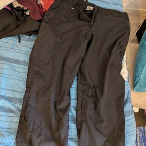 Svarta cargopants från Nytello, M - Svarta cargopants från Nytello i storlek M. Byxorna har flera stora fickor med lock och knapp, samt knappar vid bensluten för justering. Tillverkade i slitstarkt bomullstyg, perfekta för en chill och funktionell stil. Passar någon som är runt 170 cm lång.