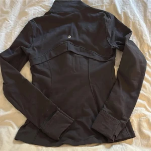 Svart träningsjacka från Lululemon - Slimmad svart träningsjacka från Lululemon med dragkedja framtill och höga muddar. Jackan har figurnära passform, ståkrage och diskret logga på ryggen. Perfekt för dig som vill ha en sportig och stilren look.