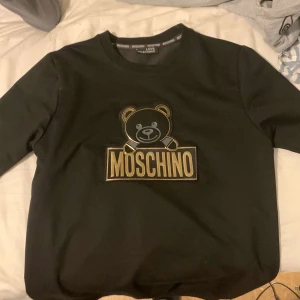 Svart Moschino sweatshirt med björn - Svart sweatshirt från Love Moschino med ikoniskt björntryck och guldig MOSCHINO-logga på bröstet. Klassisk rund halsringning och långa ärmar. Perfekt för dig som vill sticka ut med en snygg designertröja.