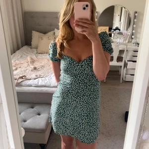 Grön blommig kortklänning - Supersöt grön kortklänning med vita små blommor, puffiga korta ärmar och söt hjärtformad urringning. Figurnära passform som framhäver midjan och ger en romantisk vibe. Perfekt för varma dagar och somriga tillfällen. Bara att skriva vid frågor. Pris kan diskuteras!