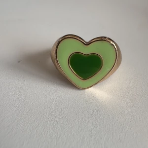 Guldig ring med grönt hjärta - Superfin ring i guldton med ett stort hjärta framtill. Hjärtat har två nyanser av grönt, en ljusare och en mörkare, vilket ger en cool kontrast. Perfekt statement-piece för dig som gillar färg och lekfulla detaljer.