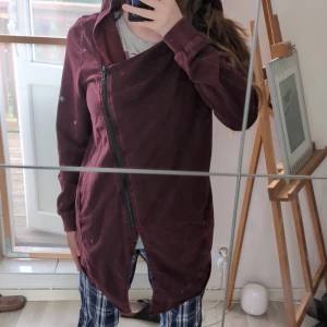 Bordeaux lång hoodiejacka med dragkedja - Säljer en bordeauxfärgad lång hoodiejacka med sned dragkedja framtill och huva. Jackan har en avslappnad passform och är tillverkad i mjukt bomullstyg. Perfekt för dig som gillar en chill och unik stil, alternativ still eller gothic feeling + den är vintage