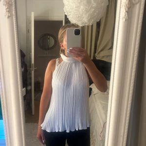 Vit plisserad ärmlös topp från Zara - Supersnygg vit plisserad topp från Zara med hög hals och knäppning i nacken. Toppen är ärmlös och har en flowy, luftig passform med snyggt fall både fram och bak. Perfekt att styla med jeans eller kjol för en trendig look.