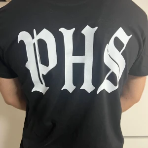 Svart PHS Support t-shirt 2023 - Svart t-shirt med stor vit PHS-tryck på ryggen och mindre tryck framtill med texten 'PHS SUPPORT 2023' och en knogjärnsdesign omgiven av lagerkrans. Klassisk passform och rund hals. Perfekt för dig som vill visa support med stil.