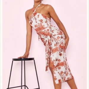 Blommig halterneckklänning med slits - Supersnygg halterneckklänning i vitt med rostbrunt blommönster. Klänningen har omlottknytning i midjan och en hög slits på sidan. Perfekt för sommarkvällar – trendig och elegant på samma gång!