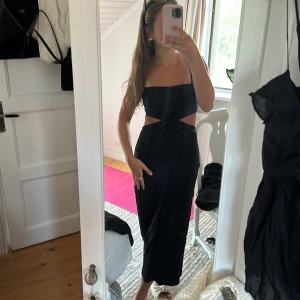 Svart långklänning med cut-outs - Säljer en svart långklänning med smala axelband och snygga cut-outs i midjan. Klänningen har en twist-detalj framtill och öppen rygg, vilket ger en trendig och stilren look. Perfekt för dig som vill sticka ut med en elegant men edgy vibe.