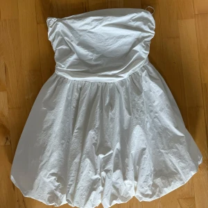 Vit axelbandslös kortklänning - Supersöt vit axelbandslös kortklänning med puffig kjol och smockad rygg. Klänningen har en ballongliknande form nertill och är perfekt för dig som vill sticka ut. Materialet känns lätt och luftigt, perfekt för varma dagar.