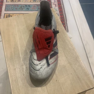 Adidas Predator fotbollssko silver/röd - Adidas Predator fotbollssko i silver med svarta och röda detaljer samt en röd plös över snörningen. Skon har dobbar för gräsplan och klassisk Predator-design med svarta ränder på sidan. Tillverkad i syntetmaterial för bra grepp och kontroll.