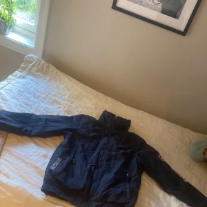 Marinblå vindjacka från Kingsland - Snygg marinblå vindjacka från Kingsland med broderad logga på ärmen och bröstet. Jackan har hög krage, dragkedja framtill och två fickor med dragkedja. Perfekt för dig som gillar ridning och vill ha en sportig look.