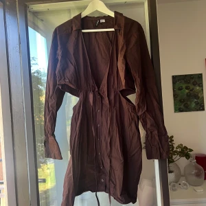Brun skjortklänning med cut-outs - Snygg brun skjortklänning med långa ärmar, knappar hela vägen framtill och coola cut-outs i midjan. Klänningen har krage och knytband i midjan för en extra detalj. Perfekt för dig som vill sticka ut med en trendig look.