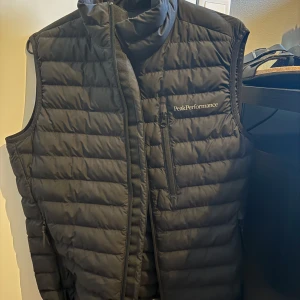 Peak Performance väst - Snygg svart dunväst från Peak Performance med quiltad design och hög krage. Västen har dragkedja framtill, bröstficka med dragkedja och två sidofickor. Perfekt för lager-på-lager och outdoor-vibe. Nypris ca 2100 ser helt ny ut