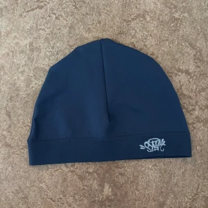 Syna World Skull Cap – Marinblå – Helt Ny & Oanvänd - Säljer en helt ny och oanvänd Syna World skull cap i marinblå färg. Tillverkad i mjuk och slitstark bomull av högsta kvalitet – otroligt skön mot huden och perfekt för alla årstider. Perfekt accessoar för att lyfta din streetwear-look. Kommer i originalförpackning med tillhörande taggar och nyckelring.