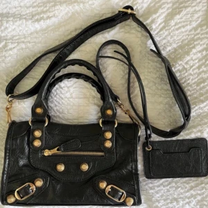Svart city bag - Snygg svart axelväska i skinn med guldfärgade nitar och spännen. Väskan har både handtag och justerbar axelrem, samt en praktisk dragkedja framtill. Inuti finns flera fack och en matchande korthållare. 