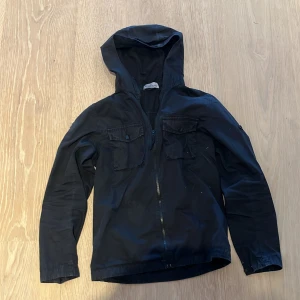 Svart vindjacka från Stone Island - Svart vindjacka från Stone Island Junior med huva, dragkedja och två bröstfickor med lock. Jackan har långa ärmar och är tillverkad i ett lätt syntetmaterial, perfekt för blåsiga dagar. Snygg och enkel design som funkar till det mesta. hål i vönster armen men lappad och lappen på höger arm saknas