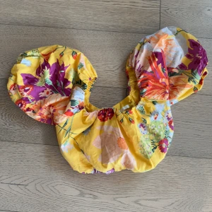 Blommig gul croppad topp H&M S - Färgglad croppad topp från H&M i storlek S med blommigt mönster i gult, lila, orange och grönt. Toppen har stora puffärmar, knytning i ryggen och elastiska detaljer för en snygg passform.