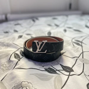 Louis Vuitton bälte  - Snyggt svart skärp från Louis Vuitton med klassiskt monogrammönster och stor LV-logga i silver på spännet. Bältet är gjort i skinn och har en brun insida. Perfekt accessoar för att lyfta din outfit med lyxig känsla.
