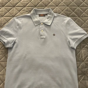 Ljusblå pikétröja från Morris - Snygg ljusblå pikétröja från Morris med klassisk krage och knappar framtill. Tröjan har korta ärmar och en liten broderad logga på bröstet. Perfekt för en clean och stilren look. Tillverkad i mjuk bomull som känns skön mot huden.