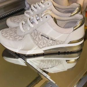 Vita sneakers från Michael Kors med silverdetaljer - Snygga vita sneakers från Michael Kors med silvriga detaljer på häl och sula. Skorna har snörning, logomönster i beige och vitt samt paneler i skinn och textil. Perfekta för dig som vill ha en clean och lyxig look med extra bling.