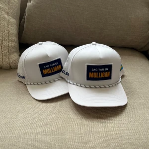 Vit keps med texten 'Jag tar en Mulligan' - Vit keps med blå och gul patch framtill där det står 'Jag tar en Mulligan'. Kepsen har perforerade sidor, böjd skärm och dekorativt snöre framtill. Svensk flagga på sidan och broderad logga. Perfekt för golf eller som statement-accessoar.