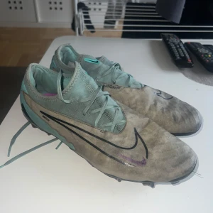 Nike Phantom GX fotbollsskor turkos - Nike Phantom GX fotbollsskor i turkos och grått med svarta och lila detaljer. Skorna har snörning, låg profil och är designade för bra grepp på planen. Swoosh-logga på sidan och Phantom GX-tryck bak. Perfekta för dig som vill sticka ut på matchen.