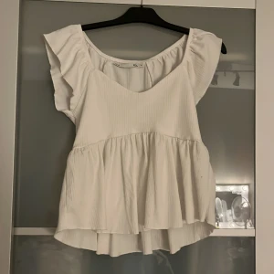 Vit volangblus från 157, storlek M - Supersöt vit blus från 157 med volangärmar och v-ringning. Toppen har en luftig, rynkad design med lätt struktur i tyget och en flowy passform som är längre baktill. Perfekt för en fräsch och somrig look!