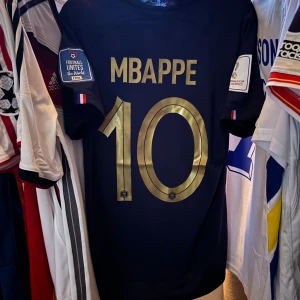 Frankrike Mbappé VM-tröja Nike blå fotbollströja 2022 - Säljer en fransk landslagströja från Nike med Mbappé och nummer 10 i guld på ryggen. Tröjan är mörkblå med guldiga detaljer, broderad tupp och två stjärnor på bröstet samt FIFA World Champions 2018-märke. Korta ärmar och lätt, ventilerande material.