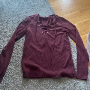 Vinröd ribbad tröja med snörning - Vinröd långärmad tröja med ribbad struktur och snygg snörning framtill vid halsen. Modellen är figurnära och passar perfekt till jeans eller kjol. Materialet är stretchigt, vilket gör den bekväm att bära hela dagen.