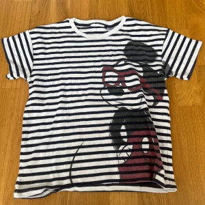 Randig t-shirt med Musse Pigg-tryck - Snygg randig t-shirt i vitt och mörkblått med ett stort tryck av Musse Pigg med röda glasögon på framsidan. T-shirten har korta ärmar och klassisk passform. Materialet känns som bomull och är mjukt mot huden. Perfekt för dig som gillar Disney och streetstyle.