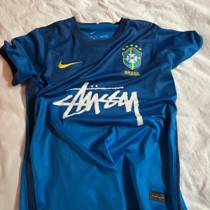 Blå Brasil Nike x Stüssy fotbollströja - Snygg blå Brasil fotbollströja från Nike i samarbete med Stüssy. Tröjan har korta ärmar, gul Nike-logga, Brasil-märke på bröstet och Stüssy-tryck i vitt. Tillverkad i lätt och ventilerande Dri-Fit-material, perfekt för träning eller match.