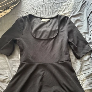 Svart tröja - Stilren svart tröja från Kappahl med fyrkantig ringning och halvlånga ärmar. Modellen är figurnära upptill och har en lätt utställd kjol. Perfekt för dig som gillar enkel och klassisk stil.