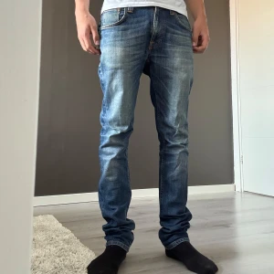 Blå jeans från Nudie Jeans, W32 L30 - Säljer ett par klassiska blå jeans från Nudie Jeans i modellen Tape Ted. Jeansen har snyggt slitna detaljer, orange sömmar och fem fickor. Tillverkade i 98% ekologisk bomull och 2% elastan för en bekväm passform.