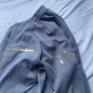 Mörkblå hoodie från Sail Racing - Mörkblå hoodie från Sail Racing med dragkedja och snygga logodetaljer på bröstet och ärmen. Jackan har en sportig look och är tillverkad i mjukt material som känns skönt mot huden. Perfekt för dig som gillar stilren och funktionell design.