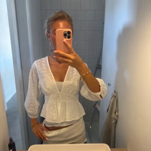 Vit blus med broderade blommor - Superfin vit blus med broderade blommor och v-ringning. Modellen har trekvartsärmar med lätt puff och knappar framtill. Blusen har en markerad midja och volang nertill, perfekt till jeans eller kjol för en fräsch look.