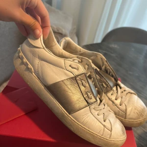 Valentino Garavani  - Storlek 40 men passar mig som har 39 i vanliga fall. Snygga sneakers från Valentino Garavani i vitt skinn med bred metallic-silver rem över vristen och coola nitar på hälen. Klassisk rund tå, platt sula och snörning framtill. Perfekta för dig som gillar lyxiga detaljer och streetstyle.