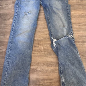 Blå lågmidjade slitna jeans  - Säljer ett par blå jeans med raka ben och slitna detaljer på låret och knät. Jeansen har klassisk femficksdesign, normal midja och är tillverkade i denim. Perfekta för en avslappnad och trendig look.